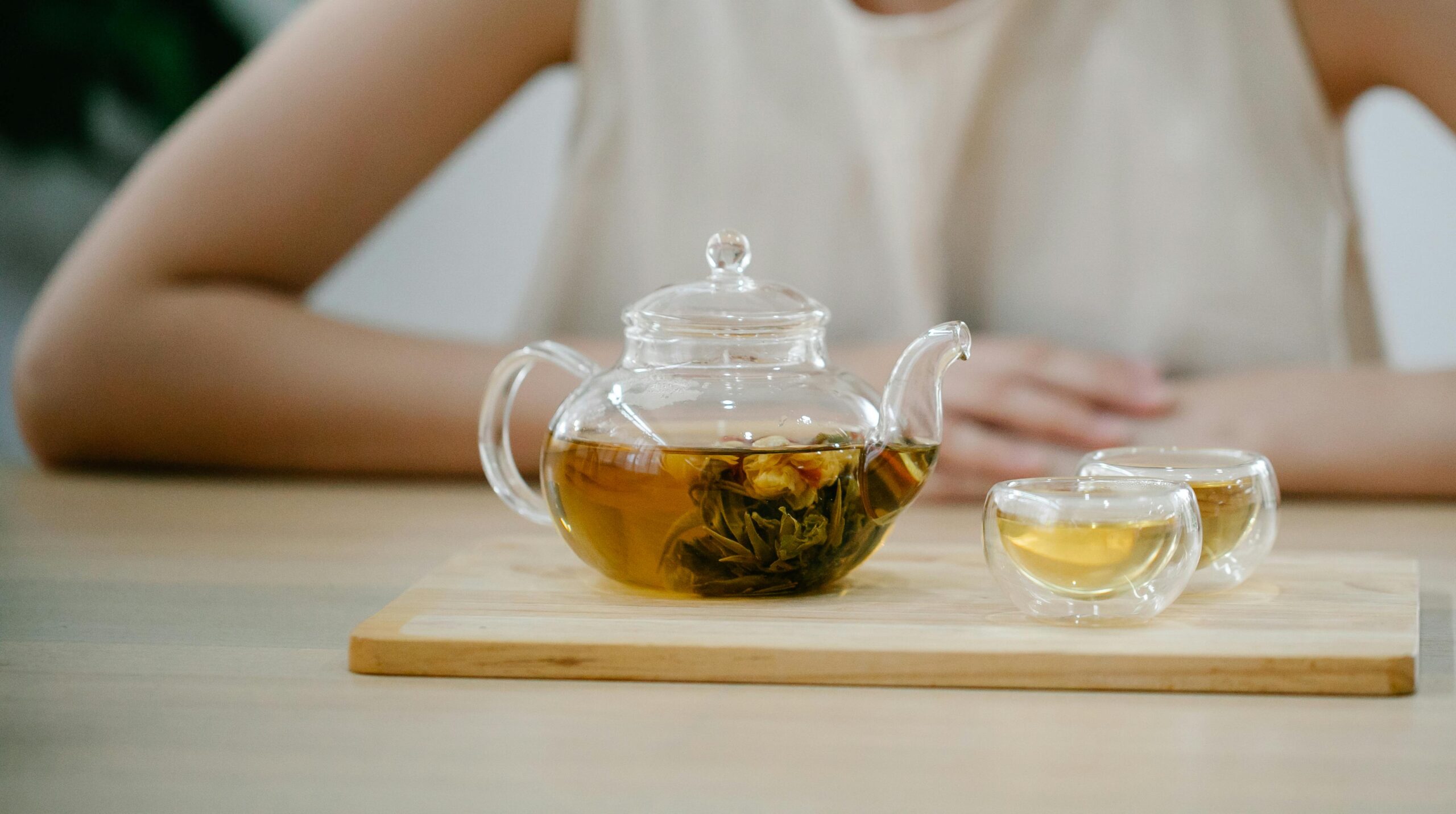 Glass teapot steeping green tea beside cups on wooden tray, creating a tranquil scene.